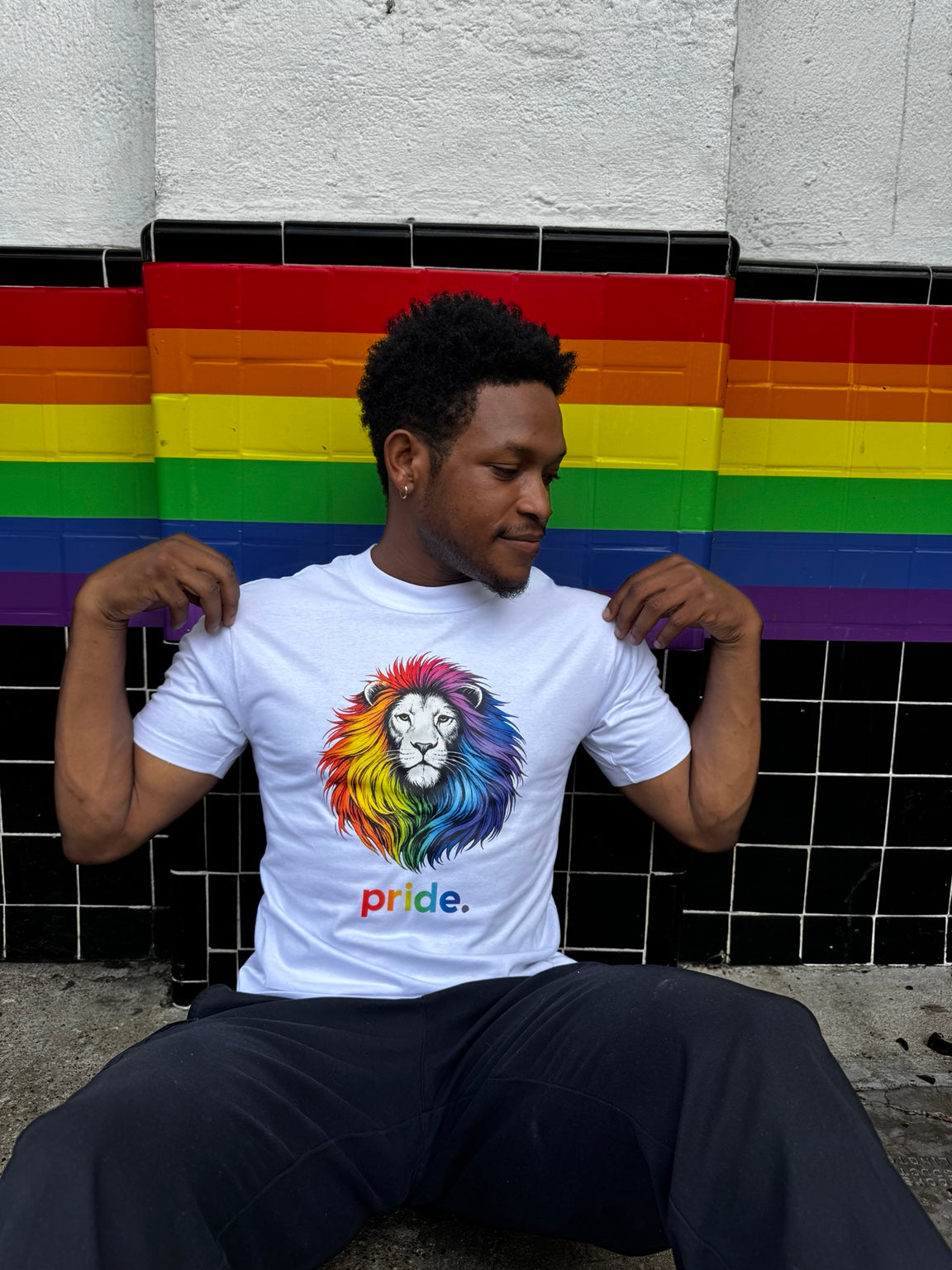 Model wearing the Poligear Pride rainbow lion graphic tee while seated against a rainbow wall
