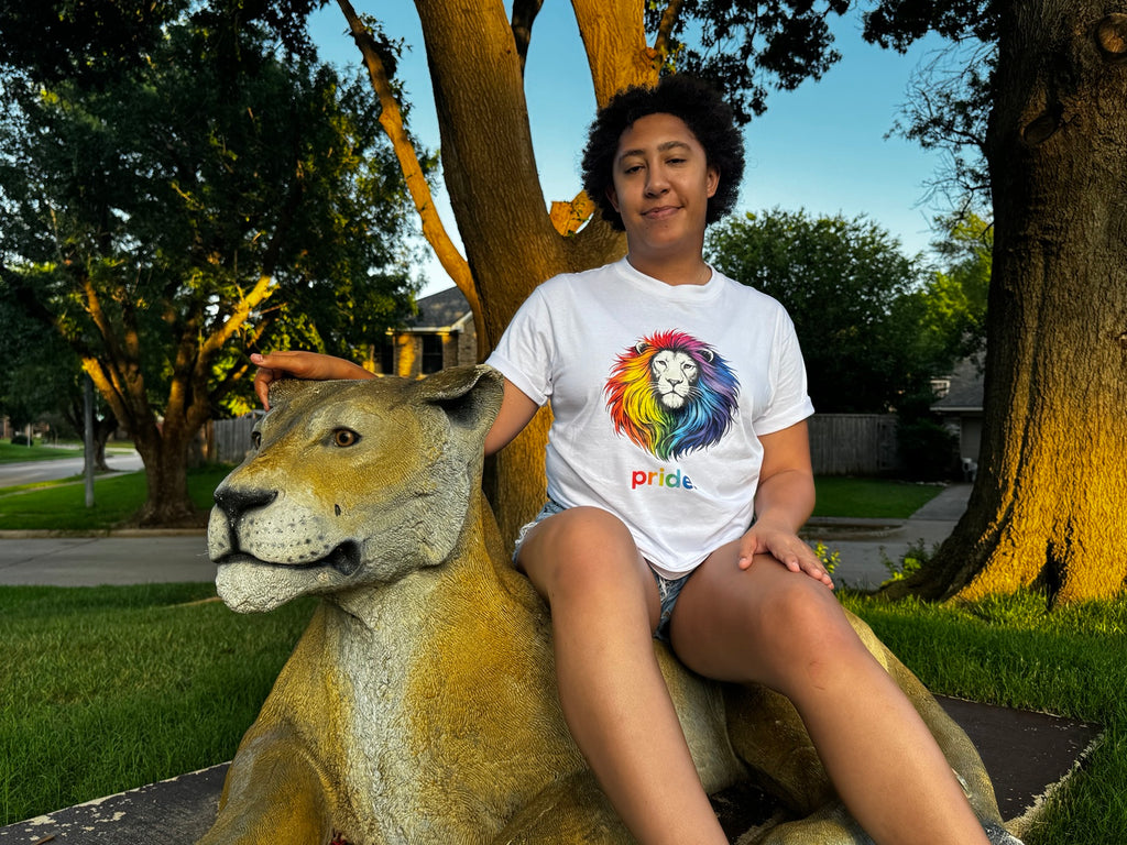 Model wearing white Pride rainbow lion tee seated on a lion statue outdoors